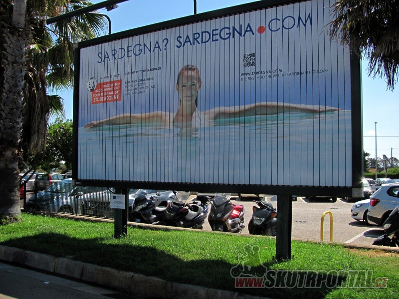 Parkoviště u letiště v Algheru, naproti vstupu. „Sardinka“ z billboardu se s námi loučí a vítá nové návštěvníky.
