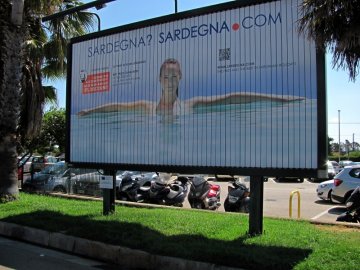 01: Parkoviště u letiště v Algheru, naproti vstupu.  „Sardinka“ z billboardu se s námi loučí a vítá nové návštěvníky.